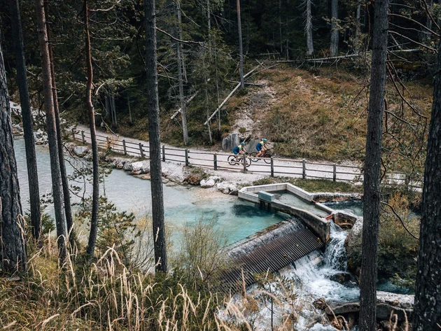 Two cyclists on forest path beside clear stream with small dam