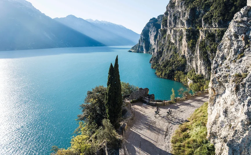 Two cyclists on mountain path by lake with steep cliffs and mountains