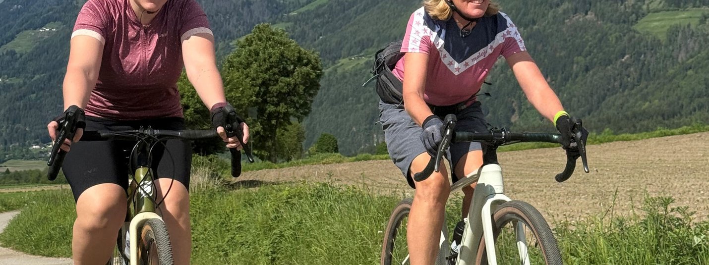 Zwei Frauen radeln auf einem Weg mit Bergen im Hintergrund