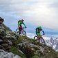 Two mountain bikers riding down a steep hill with snowy mountain peaks in the background