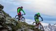 Zwei Mountainbiker fahren an einem steilen Berg mit schneebedeckten Gipfeln im Hintergrund