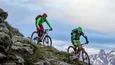 Two mountain bikers riding down a steep hill with snowy mountain peaks in the background