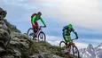 Zwei Mountainbiker fahren an einem steilen Berg mit schneebedeckten Gipfeln im Hintergrund