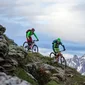 Two mountain bikers riding down a steep hill with snowy mountain peaks in the background