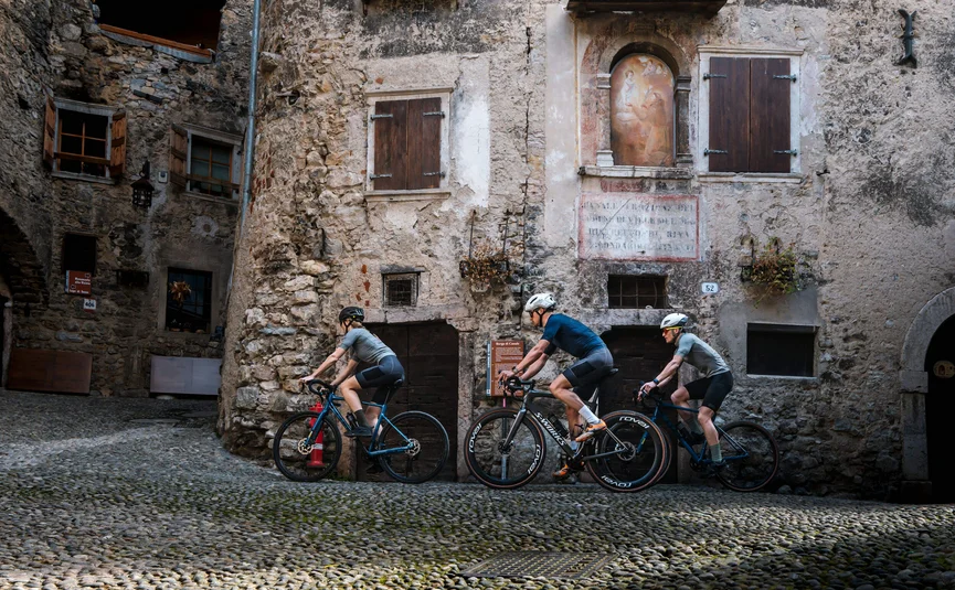 Three cyclists ride on cobblestone street past old stone buildings