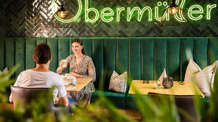 Landrefugium Obermüller – Gravelbiken im Bayerischen Wald mit Weitblick
© Stefan J. Wolf Photography Paar trinkt Kaffee in gemütlichem Café mit grünem Samtsitz und Neonbeleuchtung