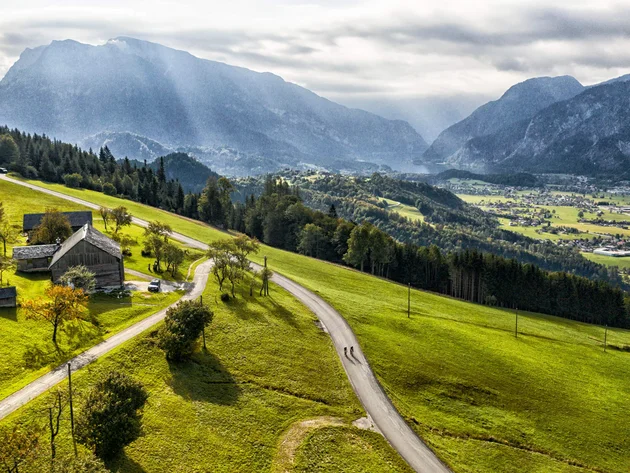 Drei Seen, eine Gravelrunde: Die 3-Seen-Tour ab Fuschl Radfahrer auf Landstraße in grüner Alpenlandschaft mit Bergen im Hintergrund