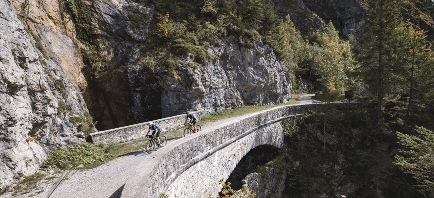 Two cyclists on a stone bridge in a mountainous landscape
