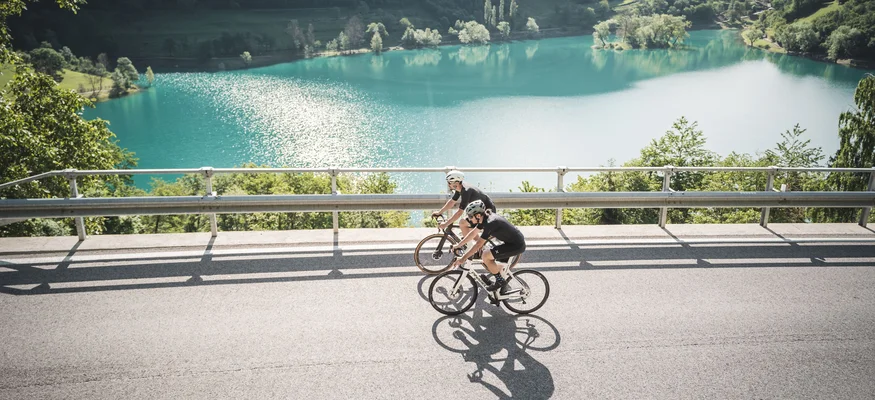 Zwei Radfahrer fahren an einem See in bergiger Landschaft vorbei