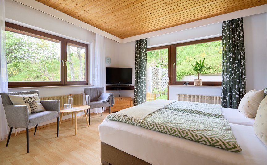 Gemütliches Schlafzimmer mit zwei Betten, Stühlen, Tisch und großen Fenstern mit Blick ins Grüne