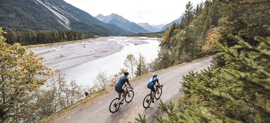 Zwei Radfahrer fahren auf einem Waldweg am Flussufer in den Bergen