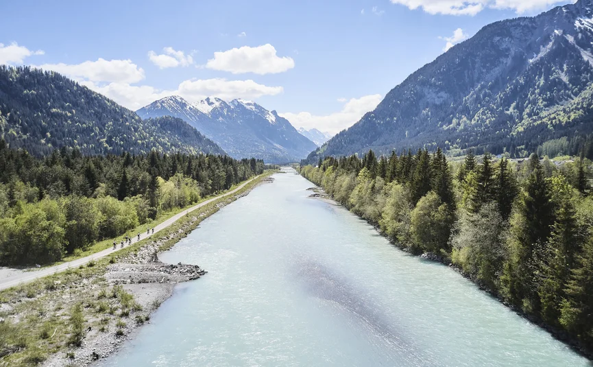 Fluss fließt durch bewaldetes Tal mit Bergen und Radweg