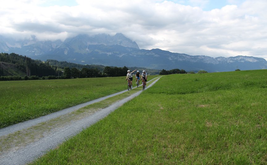 Drei Radfahrer auf einem Feldweg mit Bergen und bewölktem Himmel im Hintergrund
