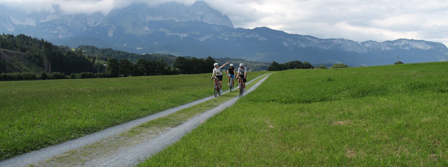 Rund um die Hohe Salve © Michael Küchl Drei Radfahrer auf einem Feldweg mit Bergen und bewölktem Himmel im Hintergrund