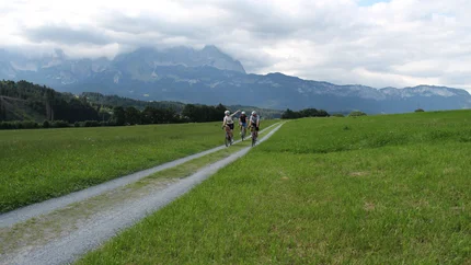 Drei Radfahrer auf einem Feldweg mit Bergen und bewölktem Himmel im Hintergrund