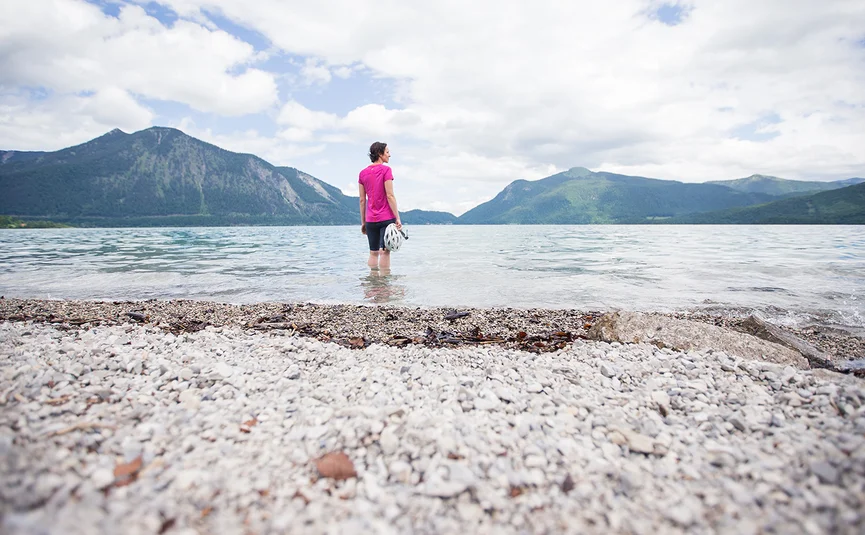 Gravelbike Urlaub in Oberbayern © Tourismus Oberbayern Frau steht im See und blickt auf Berge unter bewölktem Himmel