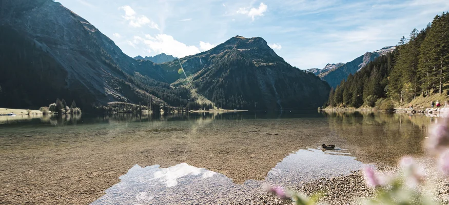 Klarer Bergsee mit steinigem Ufer und bewaldeten Bergen bei sonnigem Himmel