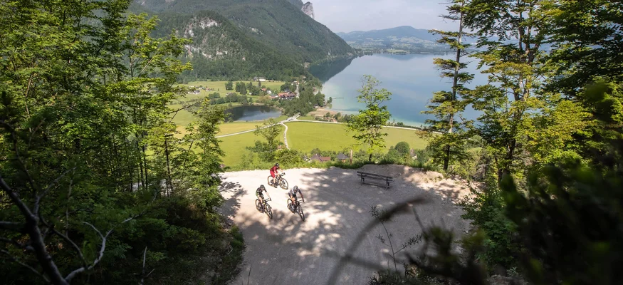 Drei Radfahrer auf einem Bergweg mit See und Bergen im Hintergrund
