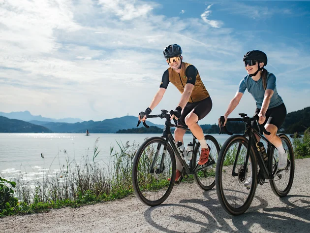 Zwei Radfahrer fahren auf einem Weg entlang eines Sees bei sonnigem Wetter