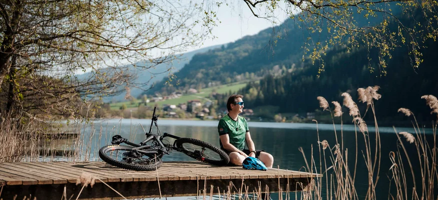 Mann sitzt auf Steg am See neben Mountainbike und genießt Bergblick.