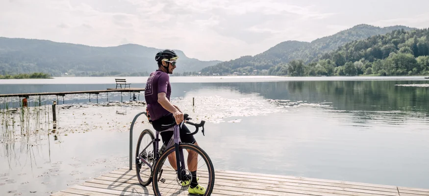 Man with bicycle sitting on a dock by a calm lake with mountains in the background