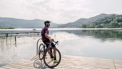 Man with bicycle sitting on a dock by a calm lake with mountains in the background