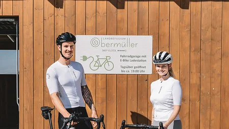 Zwei Radfahrer vor Holzgebäude mit Schild zur Fahrradgarage und E-Bike Ladestation