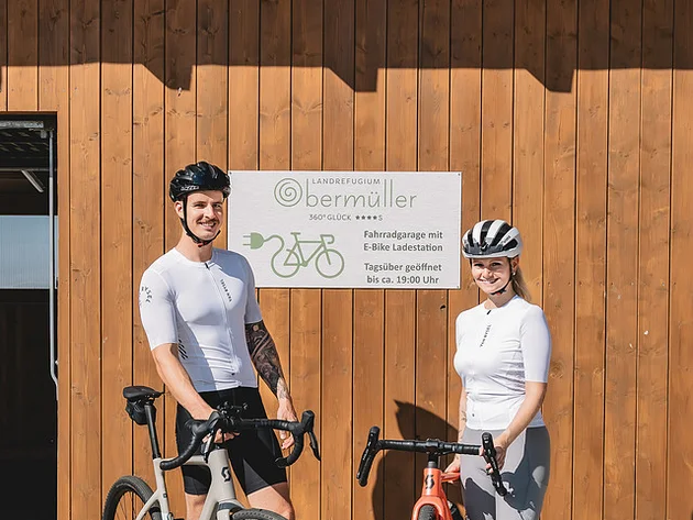Zwei Radfahrer vor Holzgebäude mit Schild zur Fahrradgarage und E-Bike Ladestation