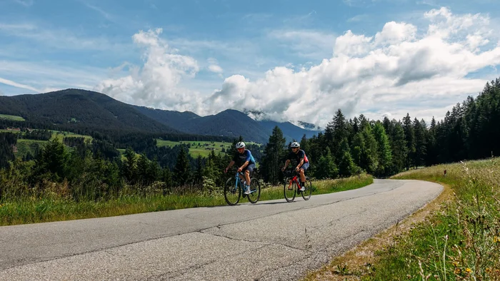 Two cyclists riding on a winding mountain road under a sunny sky