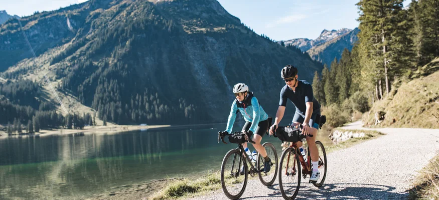 Zwei Radfahrer fahren auf einem Weg entlang eines Bergsees bei Sonnenschein