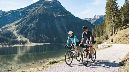 Zwei Radfahrer fahren auf einem Weg entlang eines Bergsees bei Sonnenschein