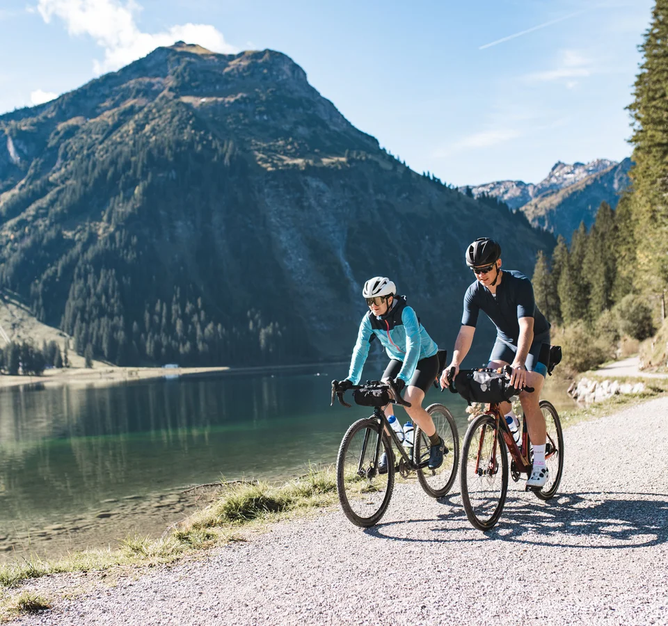 Zwei Radfahrer fahren auf einem Weg entlang eines Bergsees bei Sonnenschein