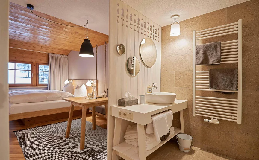 Helles Zimmer mit Bett, Holztisch und modernem Badezimmer mit zwei runden Spiegeln