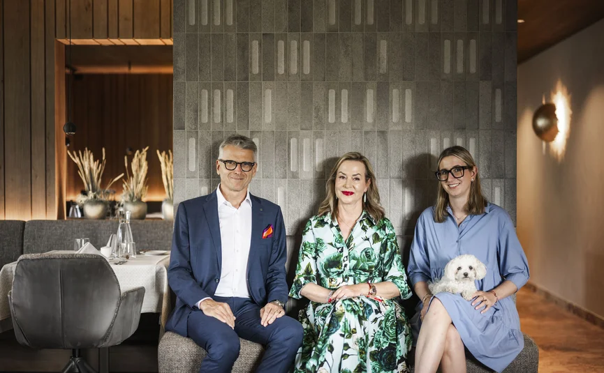 Three people sitting against a gray wall, one holding a small white dog