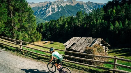 Das waren die Golden Days in Nauders © TVB-Tiroler-Oberland-Nauders-Christian-Penning - Scenic Graveltour Nauders Radfahrerin auf einem Schotterweg vor einer Berghütte und bewaldeten Alpen