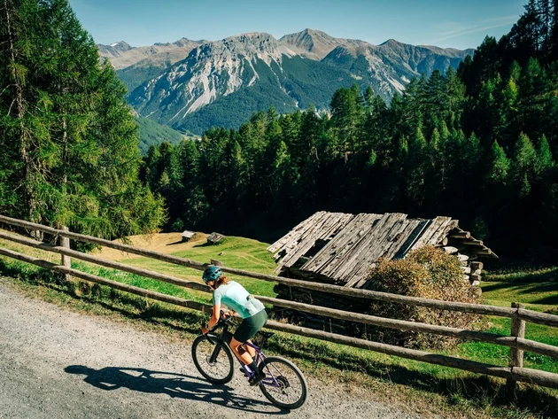 Das waren die Golden Days in Nauders © TVB-Tiroler-Oberland-Nauders-Christian-Penning - Scenic Graveltour Nauders Radfahrerin auf einem Schotterweg vor einer Berghütte und bewaldeten Alpen