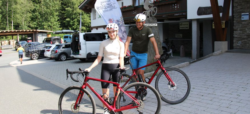 Zwei Radfahrer mit Helmen und roten Rennrädern vor einem Gebäude im Gebirge