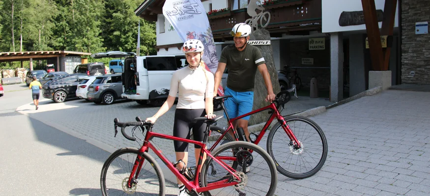 Zwei Radfahrer mit Helmen und roten Rennrädern vor einem Gebäude im Gebirge