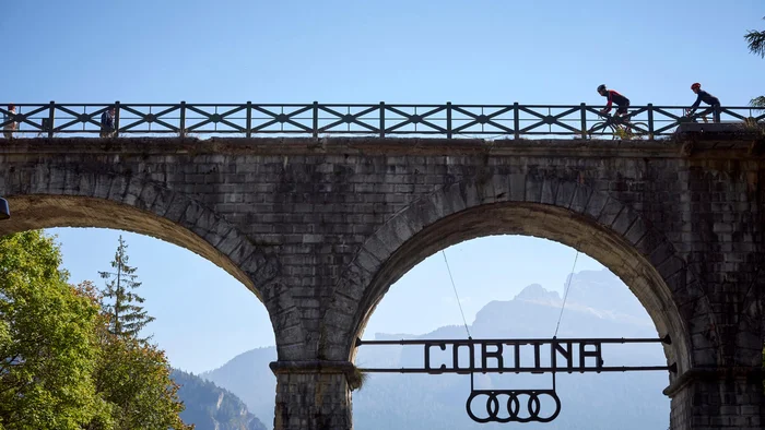 Brücke mit CORTINA-Schild und Radfahrern darüber bei klarem Himmel