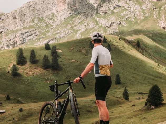 5 Gravelbike-Touren in Alta Badia, die du gefahren haben solltest © John Braynard Radfahrer mit Helm und Fahrrad steht auf grüner Hügelwiese vor Berglandschaft