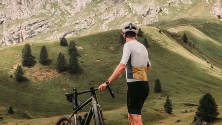 5 Gravelbike-Touren in Alta Badia, die du gefahren haben solltest © John Braynard Radfahrer mit Helm und Fahrrad steht auf grüner Hügelwiese vor Berglandschaft
