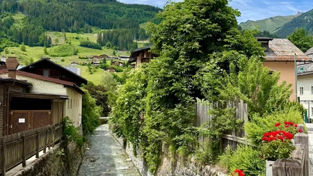 Klarer Bach fließt durch Dorf mit roten Blumen und grünen Hügeln im Hintergrund