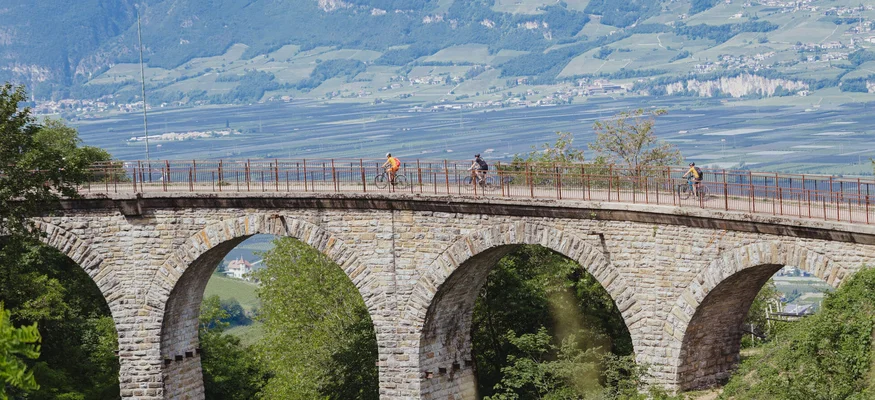Stone arch bridge with cyclists in green landscape and mountains in background