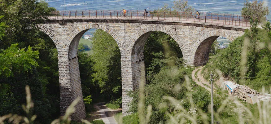 Steinbogenbrücke mit Radfahrern in grüner Landschaft und Bergen im Hintergrund