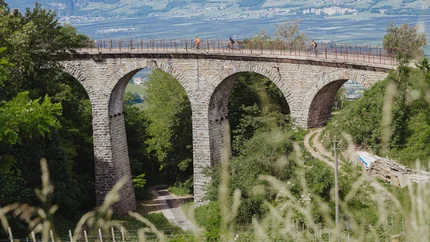 Steinbogenbrücke mit Radfahrern in grüner Landschaft und Bergen im Hintergrund
