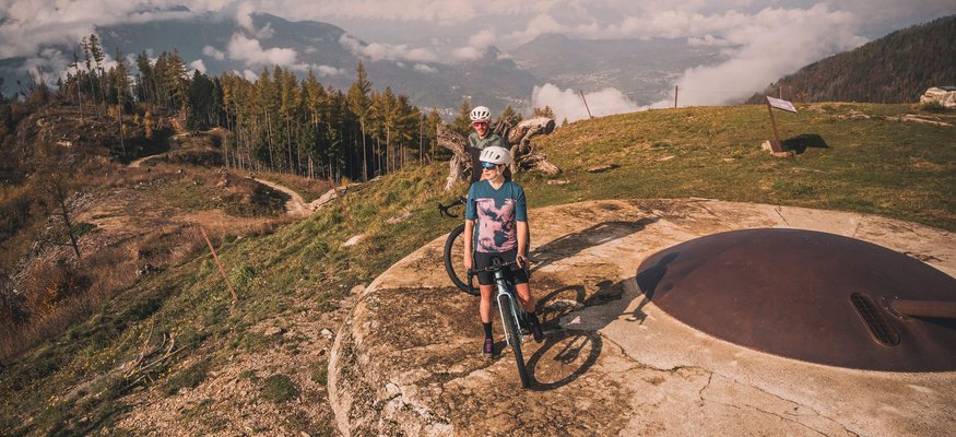Two cyclists stand on a mountain peak with fog and forest view