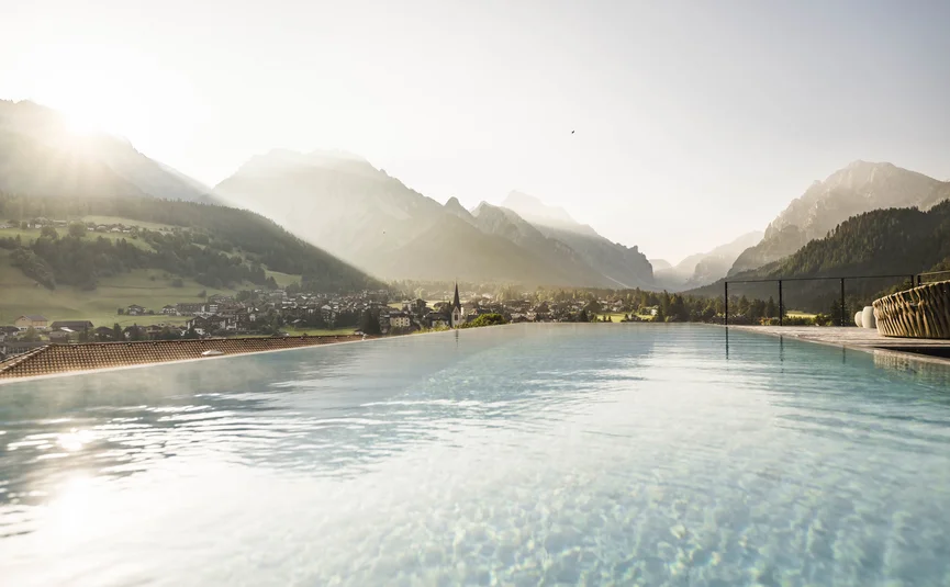 Excelsior Dolomites Life Resort ****S © Hannes Niederkofler Bergpanorama mit Dorf und Infinity-Pool im Sonnenlicht