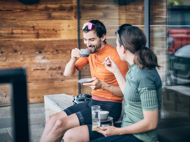 Qualitätsversprechen © Tobias Köhler Zwei Radfahrer in Sportkleidung trinken Kaffee und unterhalten sich draußen.