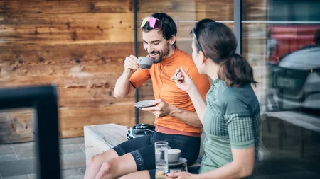 Qualitätsversprechen © Tobias Köhler Zwei Radfahrer in Sportkleidung trinken Kaffee und unterhalten sich draußen.