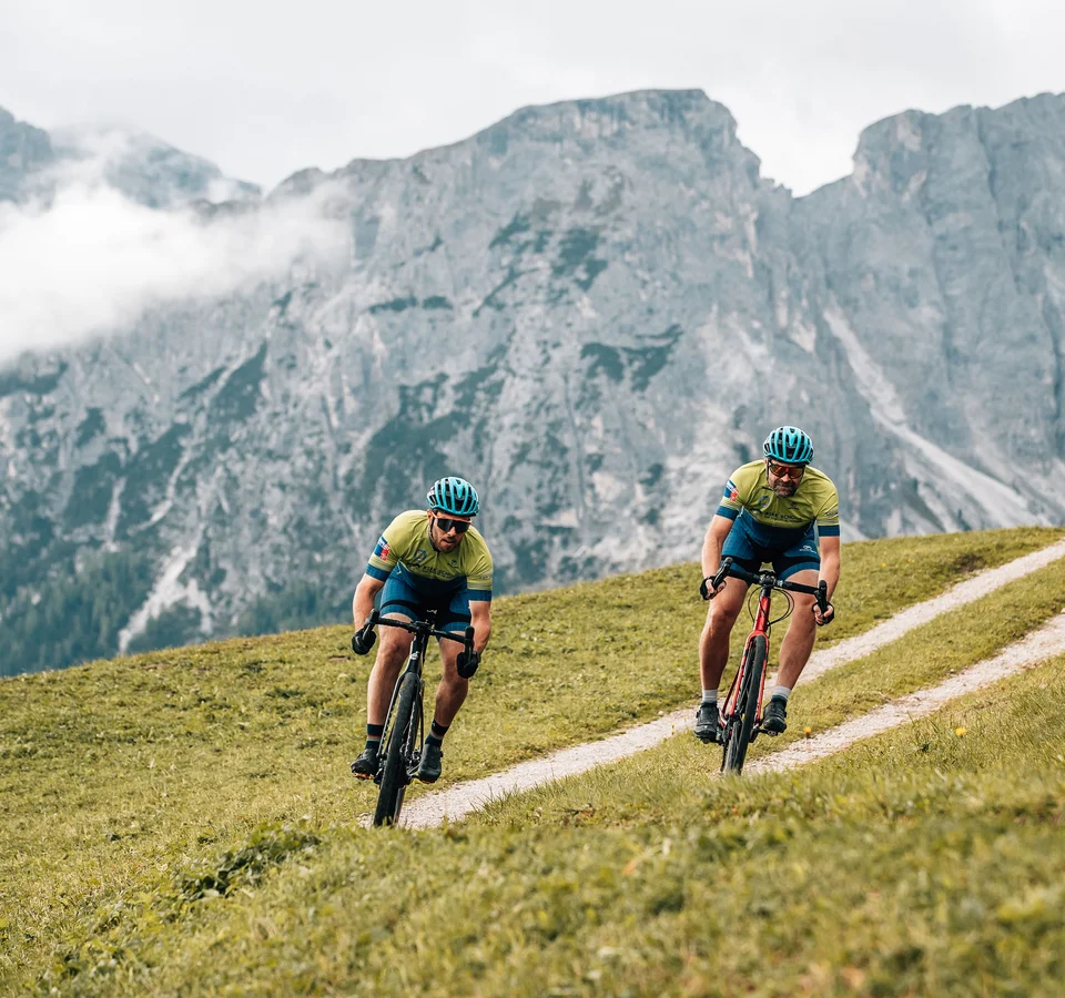 Herbst auf Schotter im Excelsior Dolomites Life Resort****s © Jens Scheibe Zwei Radfahrer fahren auf einem hügeligen Weg mit Bergen im Hintergrund